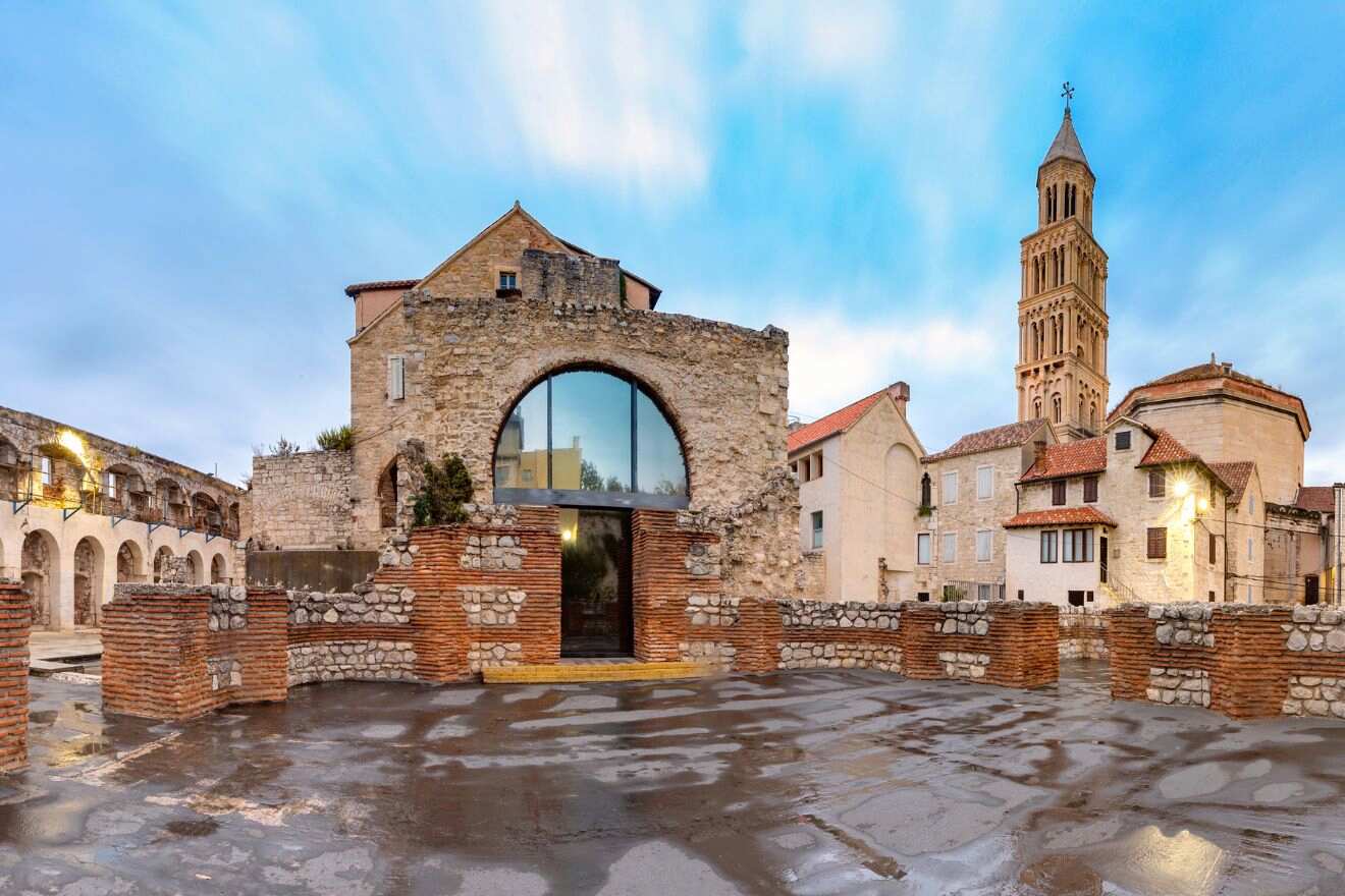 Historical ruins in Split, Croatia, with a modern glass reflection juxtaposed against ancient stone walls and a towering cathedral spire in the background under a cloudy sky