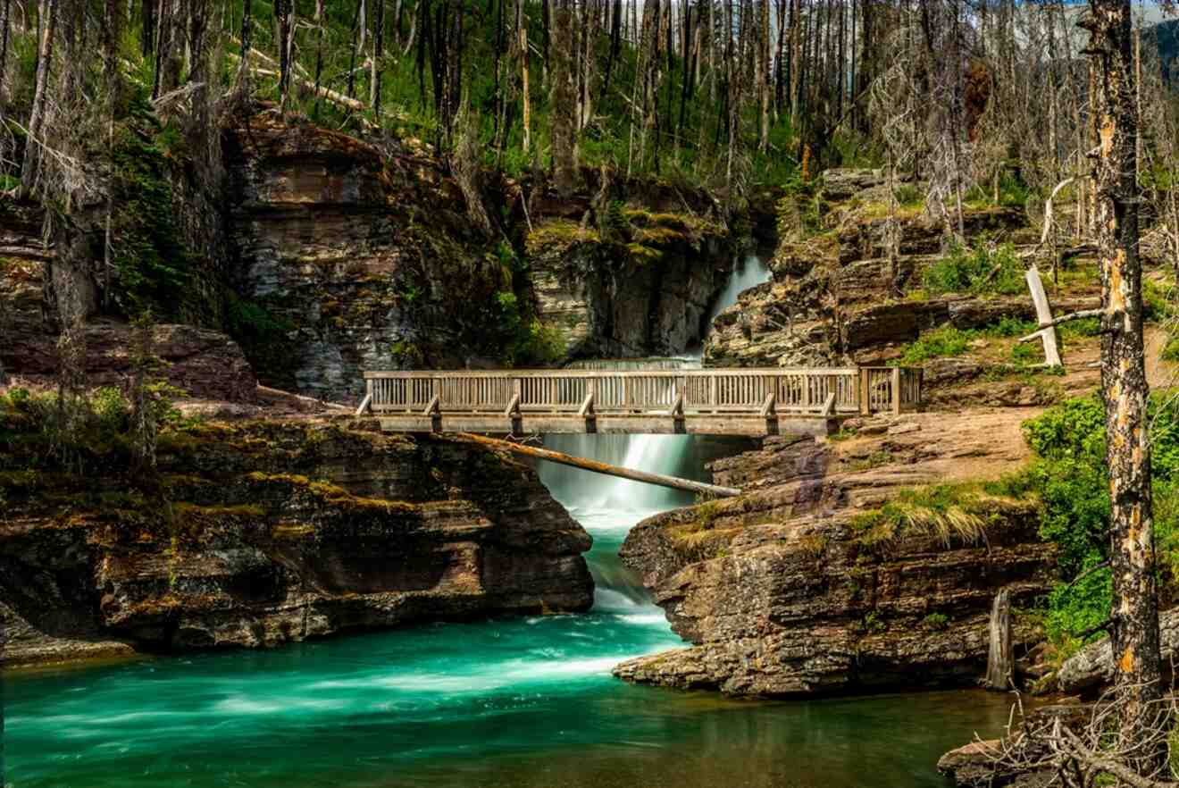 Wooden walkway bridge over a turquoise river flowing through a rocky gorge, with green trees and a waterfall in the backdrop