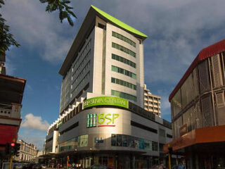 The imposing facade of BSP Suva Central, a modern multi-story building with a striking green sign, set against a backdrop of a bustling city street and blue skies