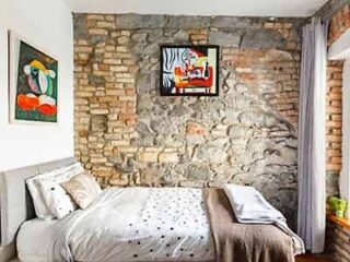 Rustic bedroom with an exposed brick wall, a comfortable bed with dotted linens, and colorful artwork hanging above.