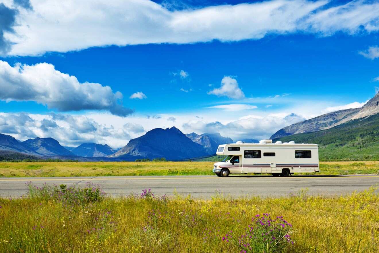 Recreational vehicle driving along a road with a view of majestic mountains and a vast field under a partly cloudy sky