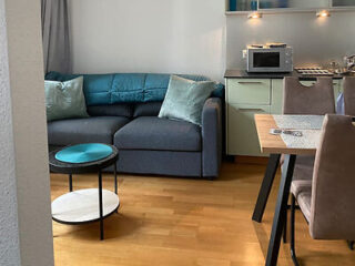 A cozy apartment living space with a blue L-shaped sofa, small round coffee table, and an open kitchenette with modern appliances and bar stools