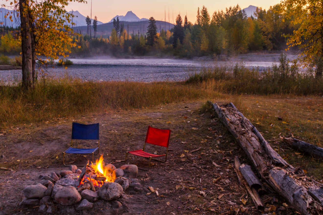 Two camping chairs around a campfire with flames, situated by a river bank with autumn trees and mountains in the distance during dusk