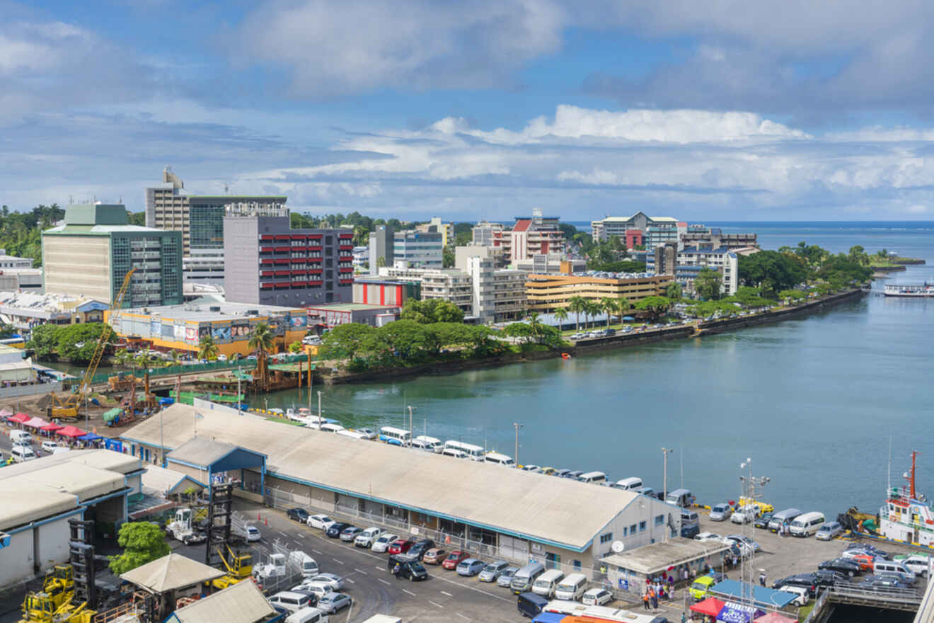 Panoramic view of a bustling coastal cityscape with modern buildings lining a calm river, a busy waterfront area with parked cars, and a vibrant blue sky dotted with white clouds overhead.