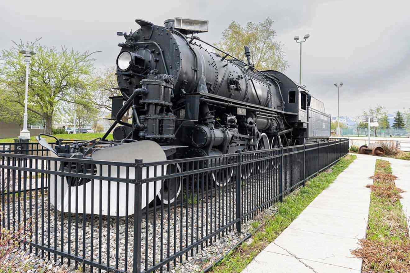 A vintage black locomotive on display, fenced off and set against a park background with trees and a cloudy sky.