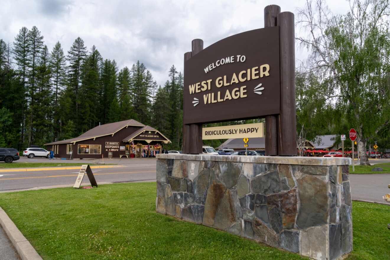 A sign reading 'Welcome to West Glacier Village' with cheerful messages, in front of a gift shop and parking lot, under a cloudy sky