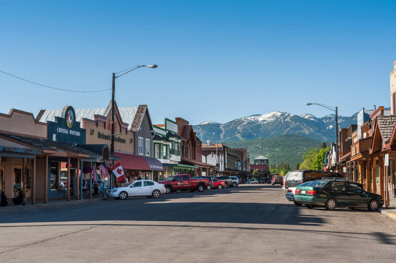 Quaint mountain town street lined with local businesses and cars, with Canadian flags and a view of snow-capped mountains in the distance