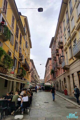 A quaint, narrow street in Milan lined with colorful buildings and filled with outdoor café seating, pedestrians, and a cobblestone walkway