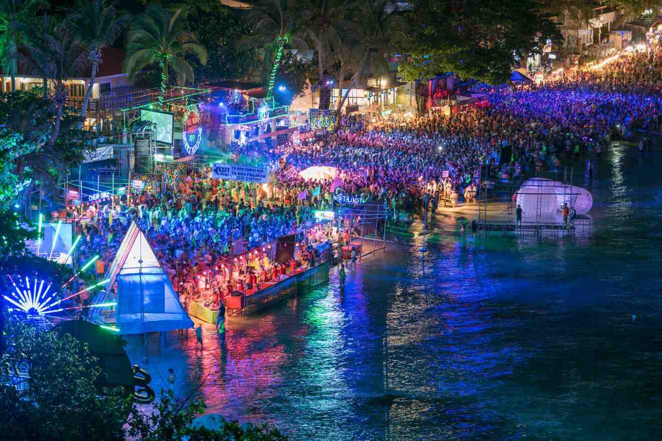 A nighttime shot of a lively beach party in Haad Rin, Koh Phangan with a dense crowd illuminated by vibrant lights, reflecting on the water with boats docked nearby.