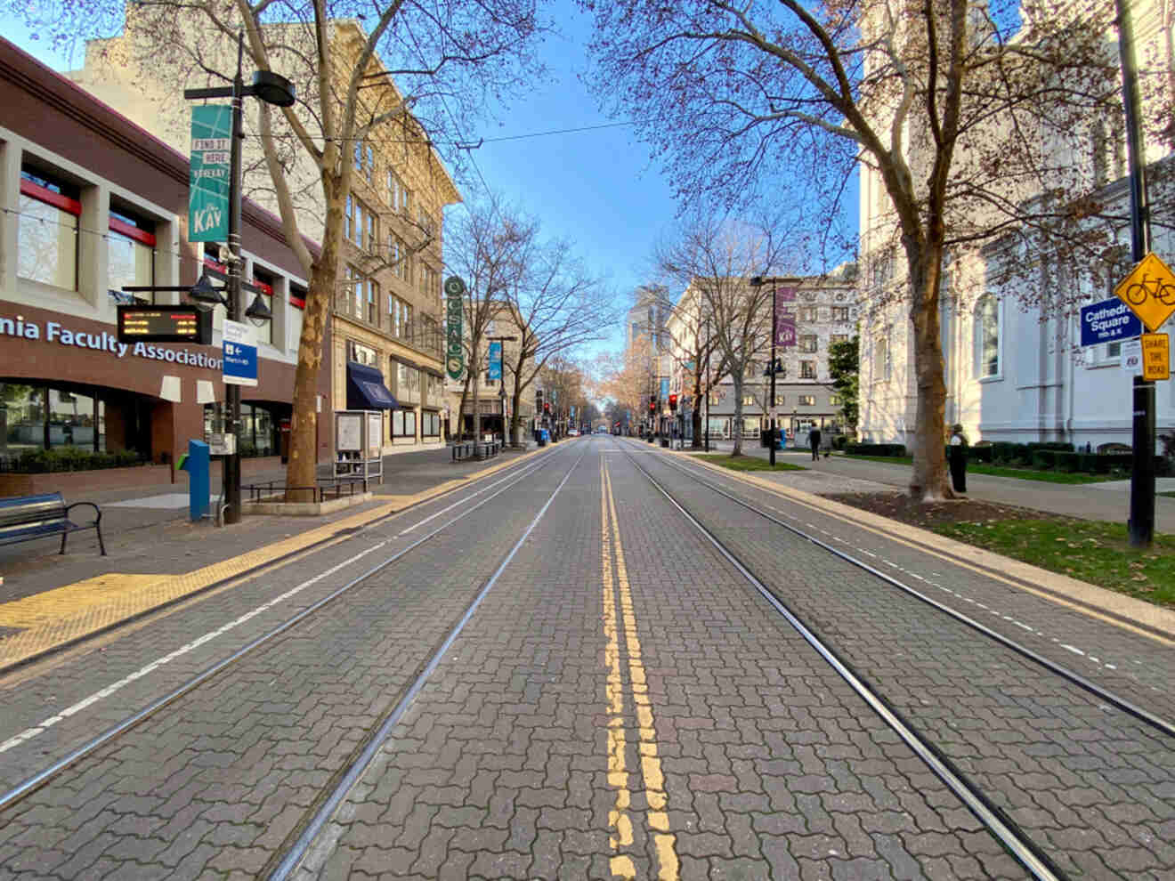 a quiet, tree-lined city street in Midtown Sacramento with tram tracks running down the center. Storefronts and historical buildings flank one side, while a clear blue sky adds to the serene urban landscape