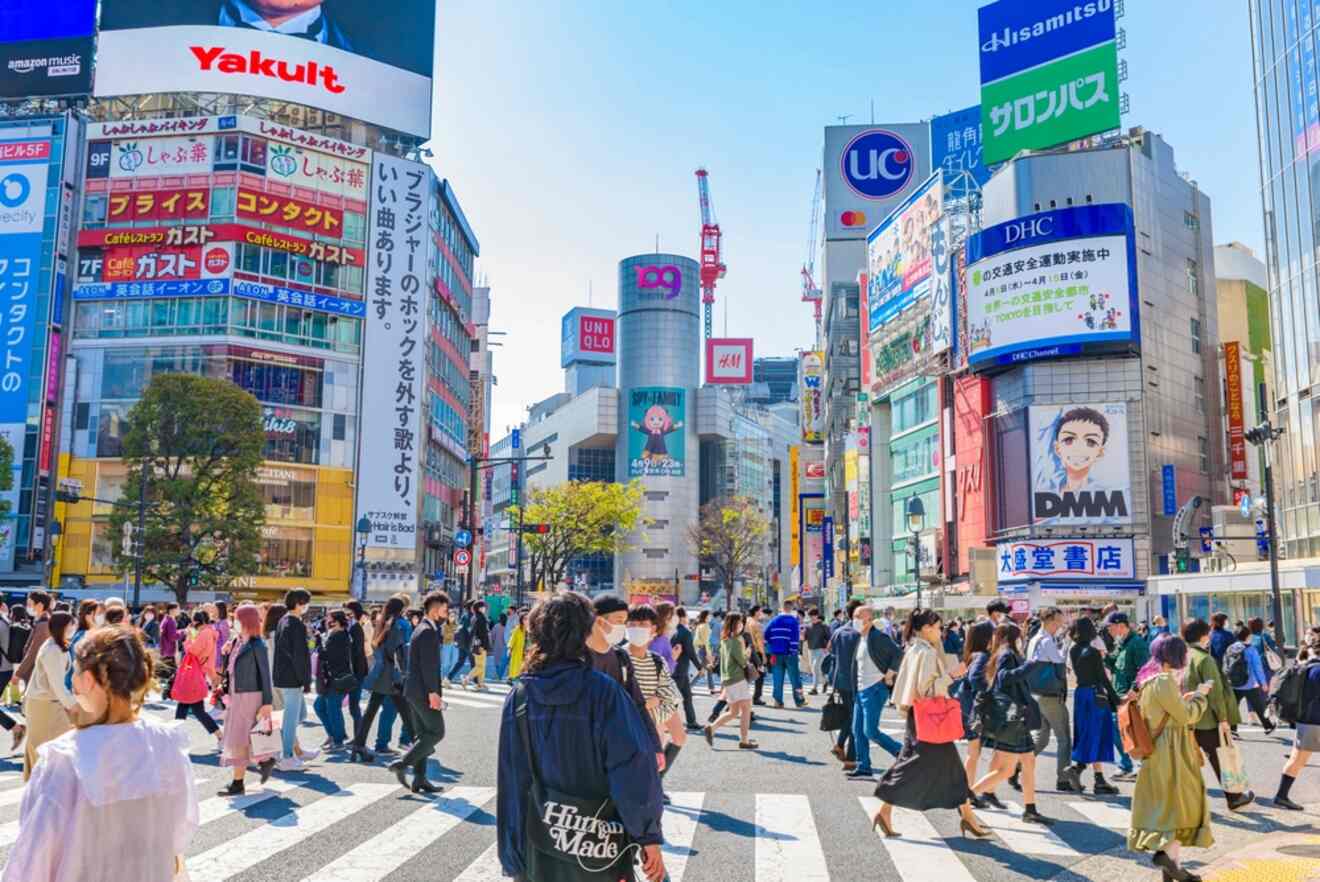 Daytime scene of a crowded Shibuya Crossing with pedestrians and vibrant advertising billboards under a clear blue sky