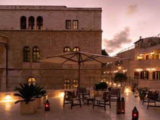 Evening view of a hotel courtyard with lit lanterns, outdoor seating under umbrellas, and a historic stone building in the background.