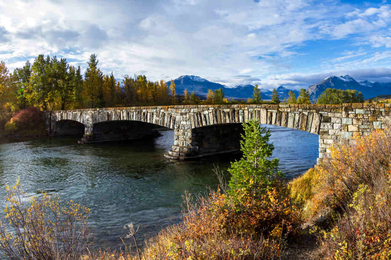Stone bridge spanning a river surrounded by fall foliage and mountain scenery under a partly cloudy sky