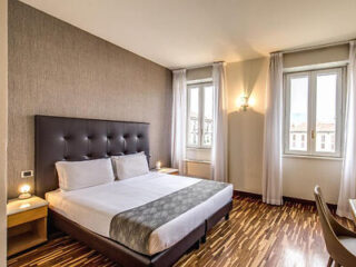 A neat and modern hotel room with a large bed featuring a tufted headboard, wood flooring, gray walls, and white drapery, offering a warm and inviting atmosphere