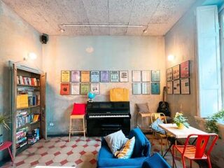 A cozy hostel common room with eclectic decor, featuring a blue sofa, a red piano, colorful framed art on the walls, and a bookshelf filled with books