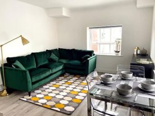 A modern living room with a plush green velvet sofa, patterned rug, and a glass dining table set for two