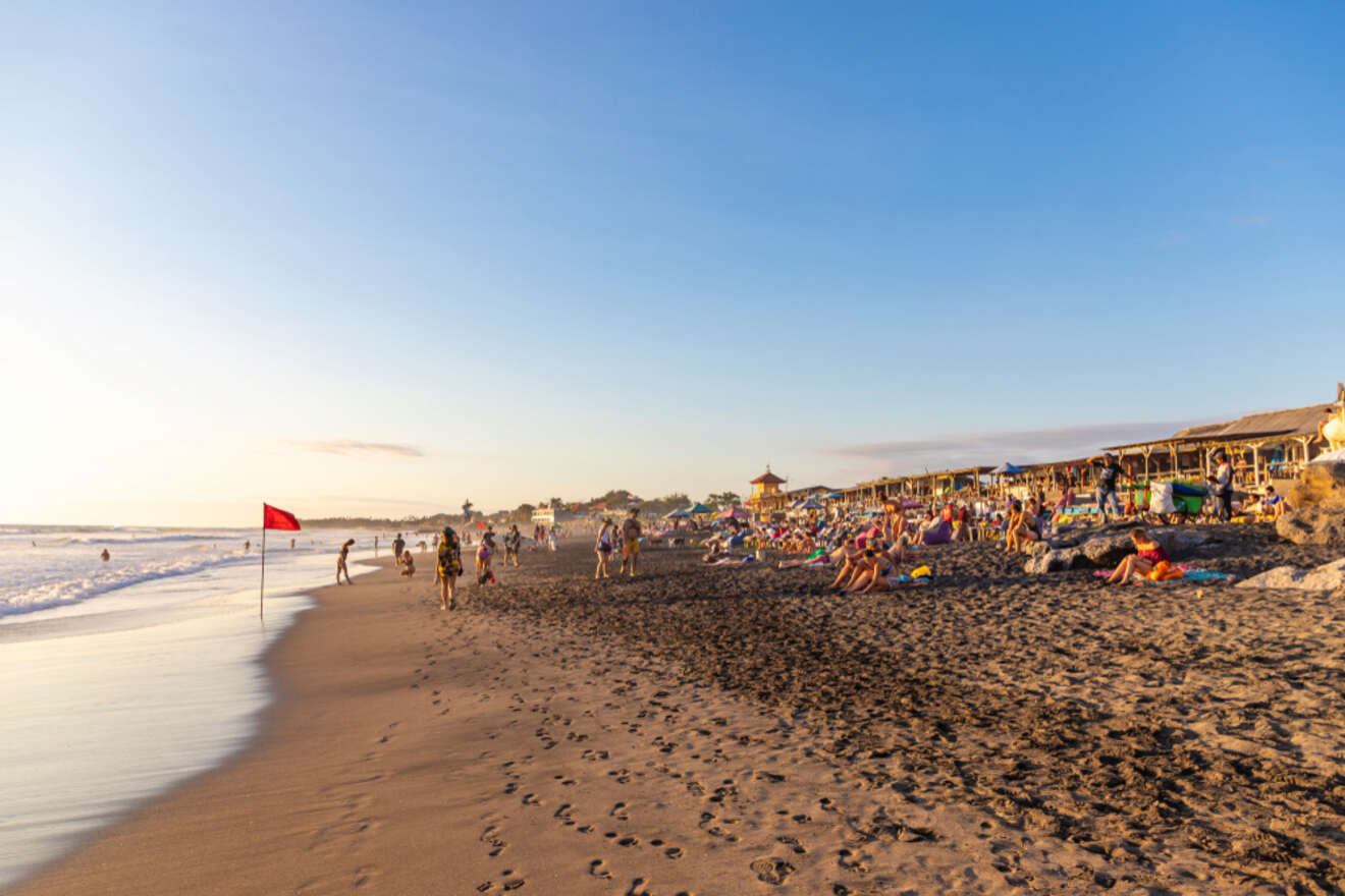 A bustling beach scene at sunset with a red flag fluttering in the wind, visitors walking along the shoreline, and beachgoers lounging on the sand under the warm glow of the evening sky.