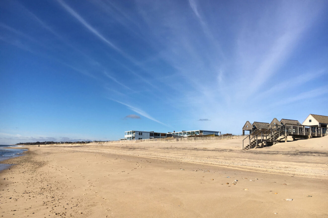 A wide sandy beach in Amagansett with beach houses in the distance under a blue sky with wispy clouds, representing a family-friendly destination in the Hamptons