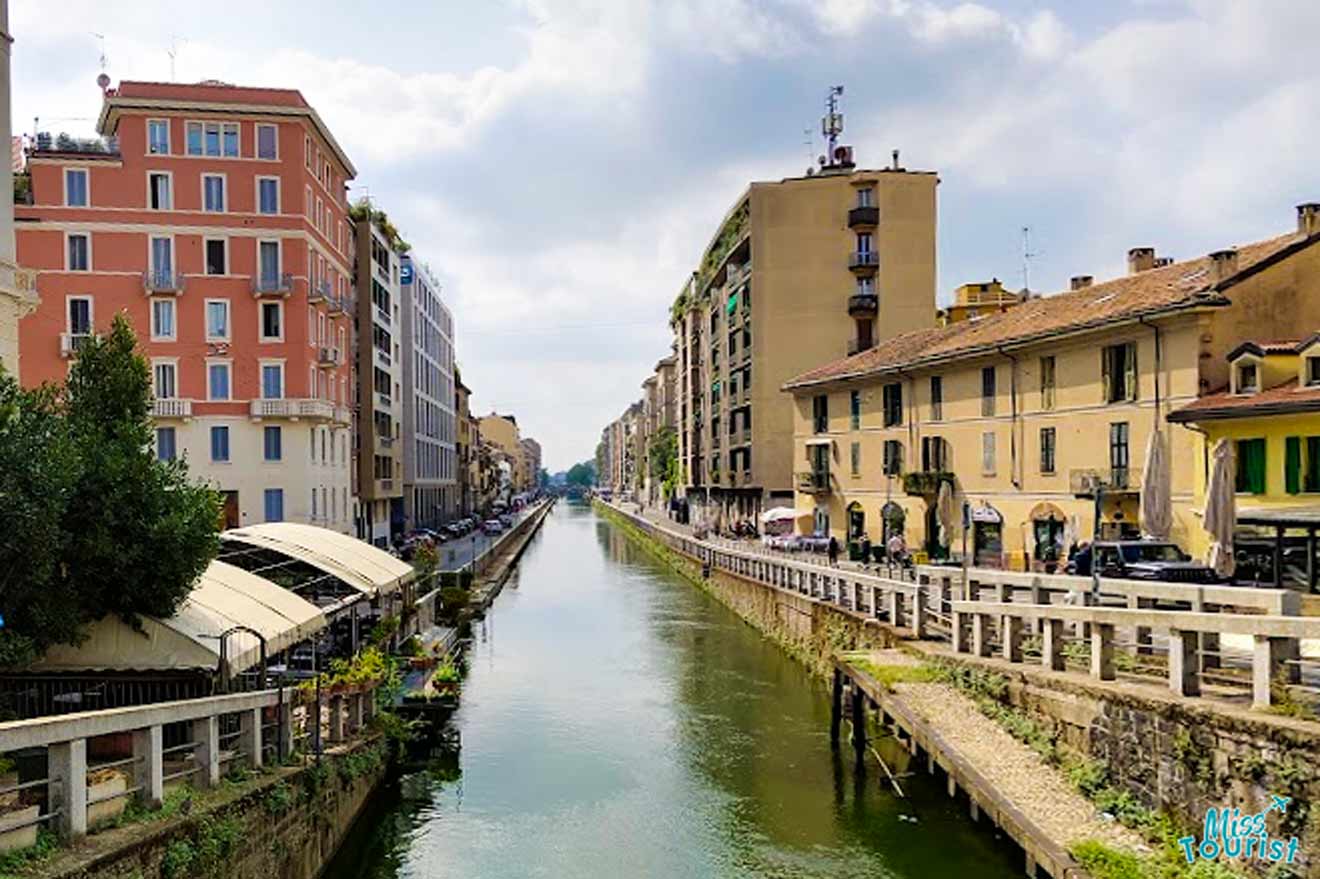 A serene canal scene in Milan's Navigli district, with a tree-lined waterway, restaurants along the banks, and traditional buildings under a partly cloudy sky