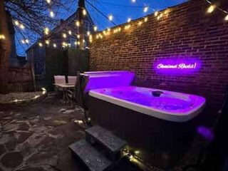 An outdoor hot tub in an intimate courtyard setting with brick walls, string lights, and a neon sign, creating a relaxing urban oasis.