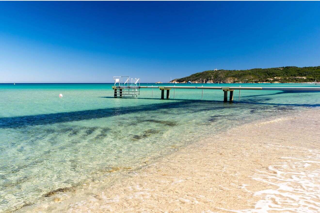 A tranquil beach scene in Saint-Tropez with crystal clear waters gently lapping the shore, a small pier extending into the serene sea, all under a clear blue sky