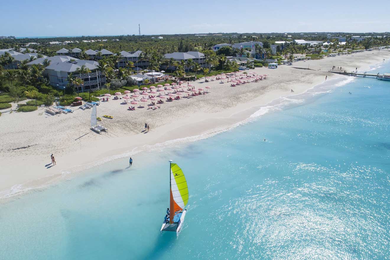 Aerial view of a luxurious beachside resort in Leeward Settlement with pink umbrellas lining the white sandy beach and a single colorful sailboat on the clear turquoise water