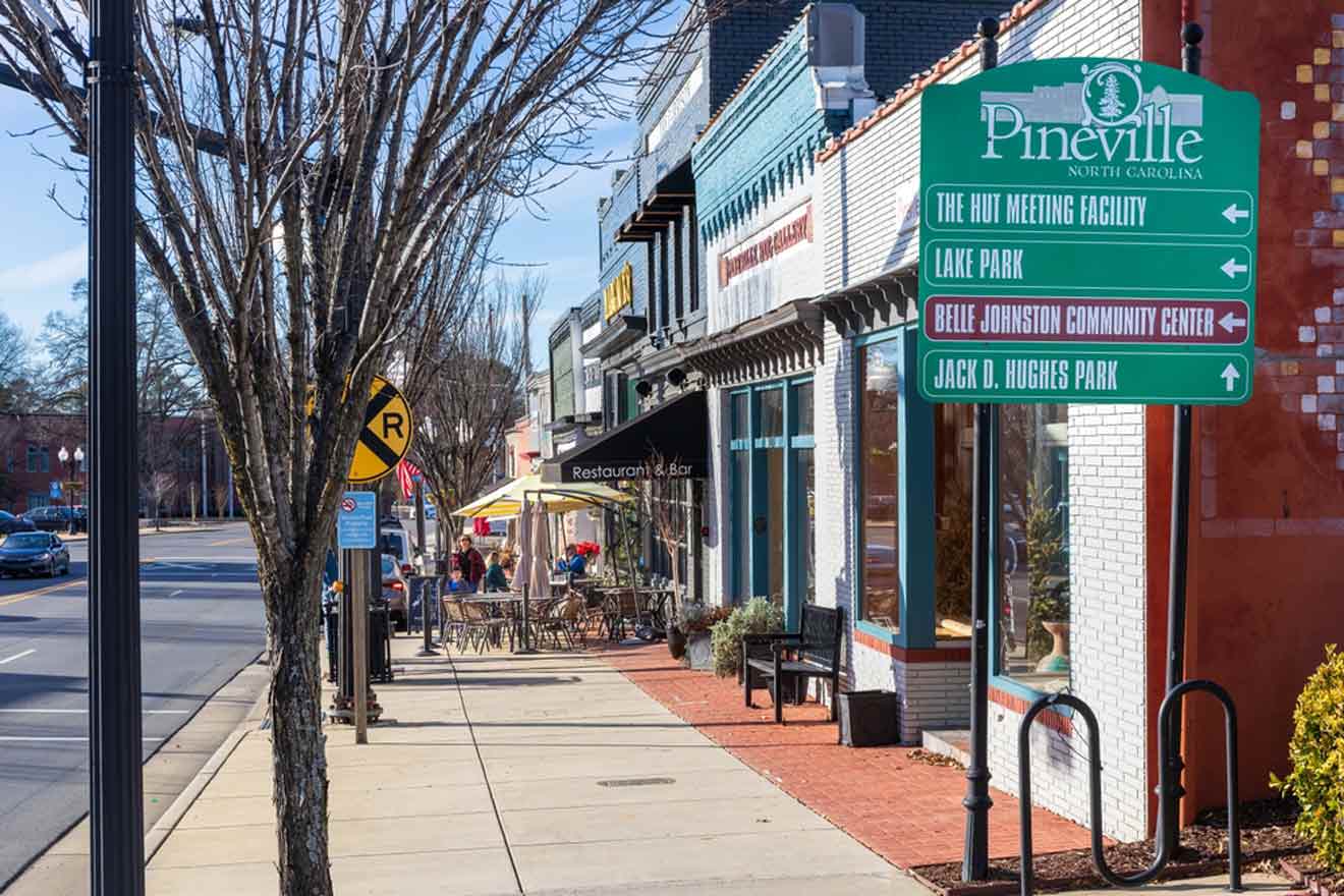 Sunny day view of a bustling street in Pineville, North Carolina, with outdoor seating at a restaurant, a variety of storefronts, and a directional sign pointing to local attractions such as The Hut Meeting Facility, Lake Park, Belle Johnston Community Center, and Jack D. Hughes Park