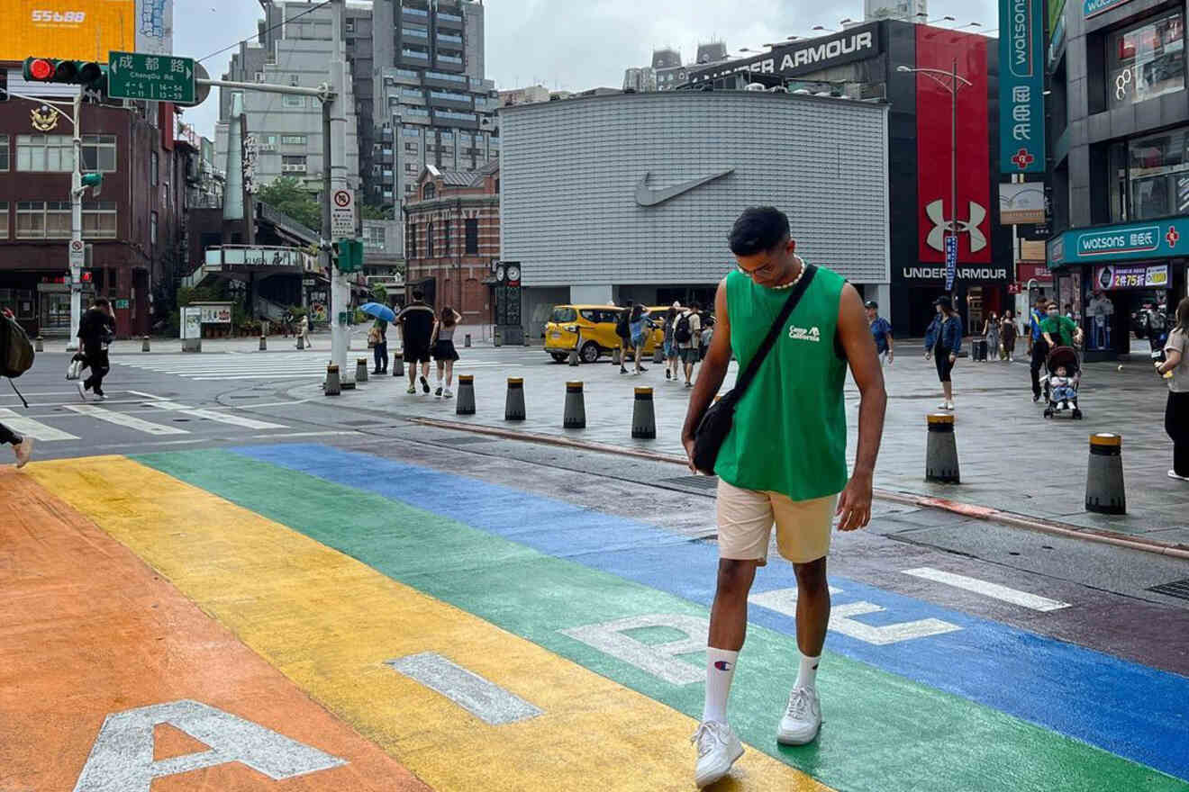 A street crossing in Ximending, Taipei's bustling shopping district, with a rainbow-painted crosswalk and a young person in casual summer clothing.