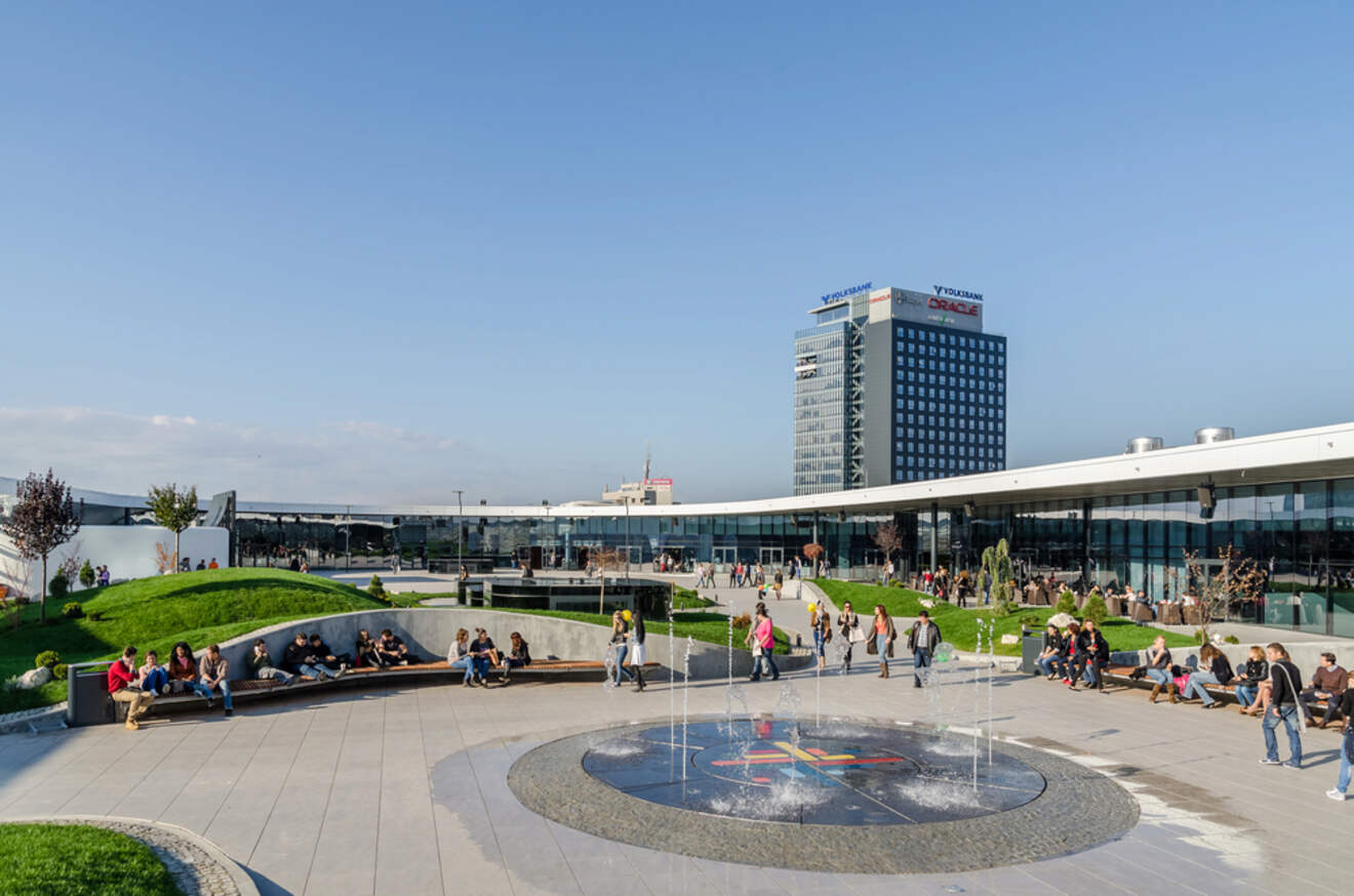 A modern urban park with people sitting around a fountain, with a tall office building in the background.