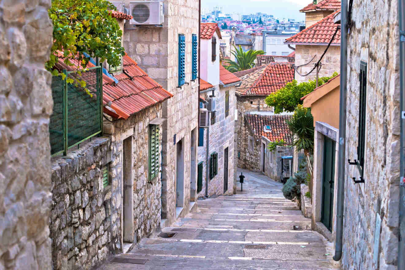 Quaint stone-paved alleyway descending between traditional Croatian houses with shutters, leading towards a cityscape under a partly cloudy sky in Split
