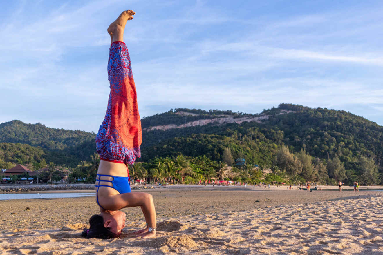 A woman performing a headstand on a sandy beach in Srithanu, Koh Phangan with the sun setting over a tree-lined coast in the background