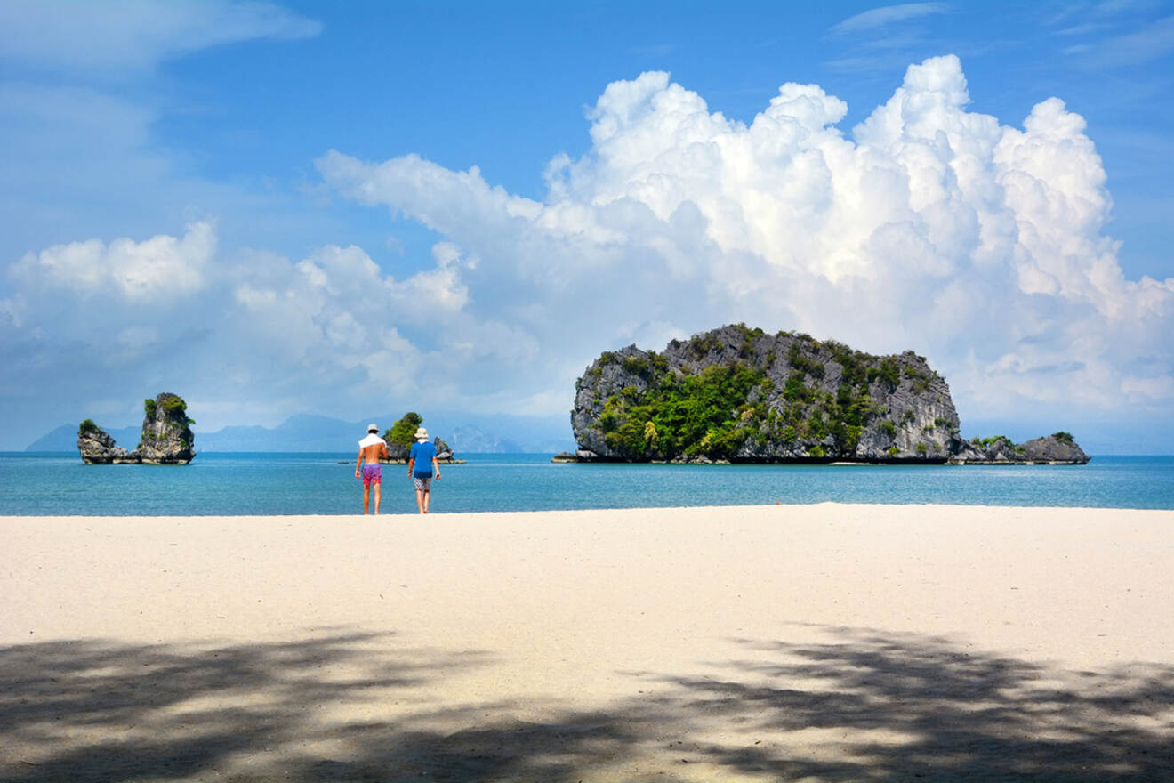 Two tourists walking towards unique limestone formations in a calm sea in Langkawi under a blue sky with dramatic clouds.