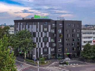 The Ibis Styles Hotel, a contemporary building with a dark facade highlighted by square windows, stands prominently at a city street corner, under a sky with soft clouds.