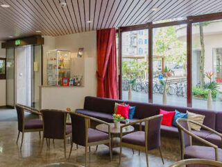 Hotel lobby with comfortable seating, purple sofas with red cushions, round tables, under a wooden slatted ceiling by large windows overlooking the street
