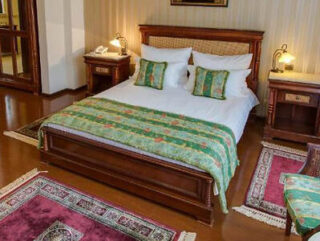 An elegant hotel room featuring classic furniture, a comfortable bed with green and gold bedding, wooden floors, and decorative elements