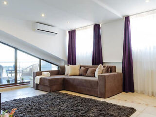 A spacious and well-lit penthouse living room with a large, plush brown sectional sofa, purple drapes, and a balcony offering a view of the city skyline