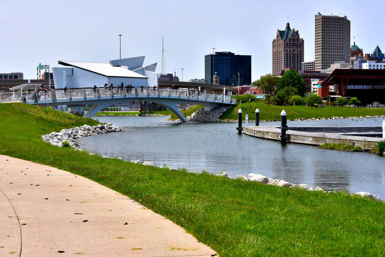 A pedestrian bridge over the Menomonee River in the Walker's Point neighborhood with the Milwaukee skyline in the background, showcasing a mix of modern and historical architecture on a clear day