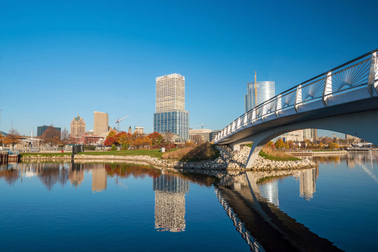 A serene morning view of the Milwaukee skyline reflected in the calm waters, with a modern pedestrian bridge in the foreground, set against a clear blue sky