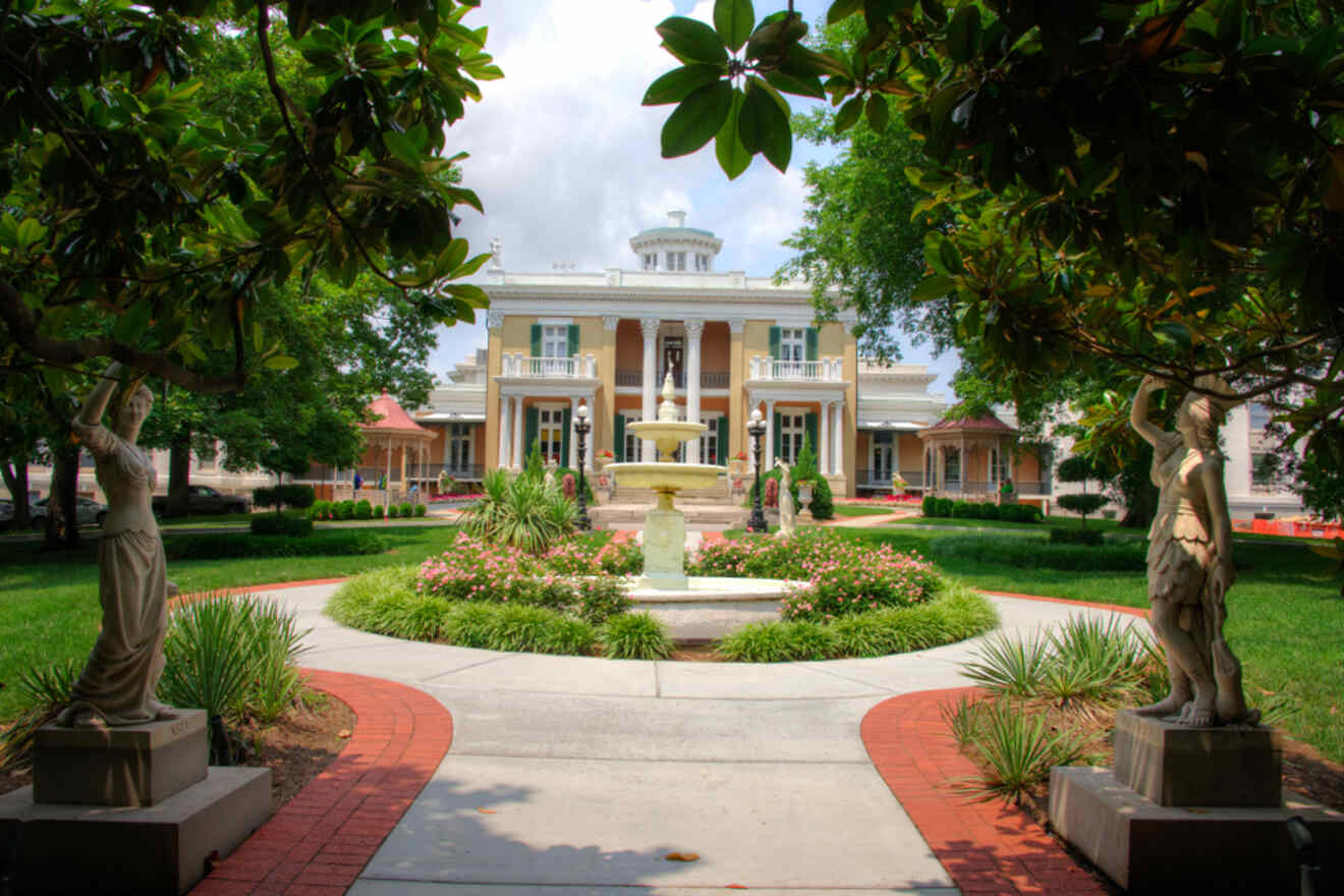 Historic southern mansion with a grand white facade, surrounded by manicured gardens and classical statues, under a clear blue sky
