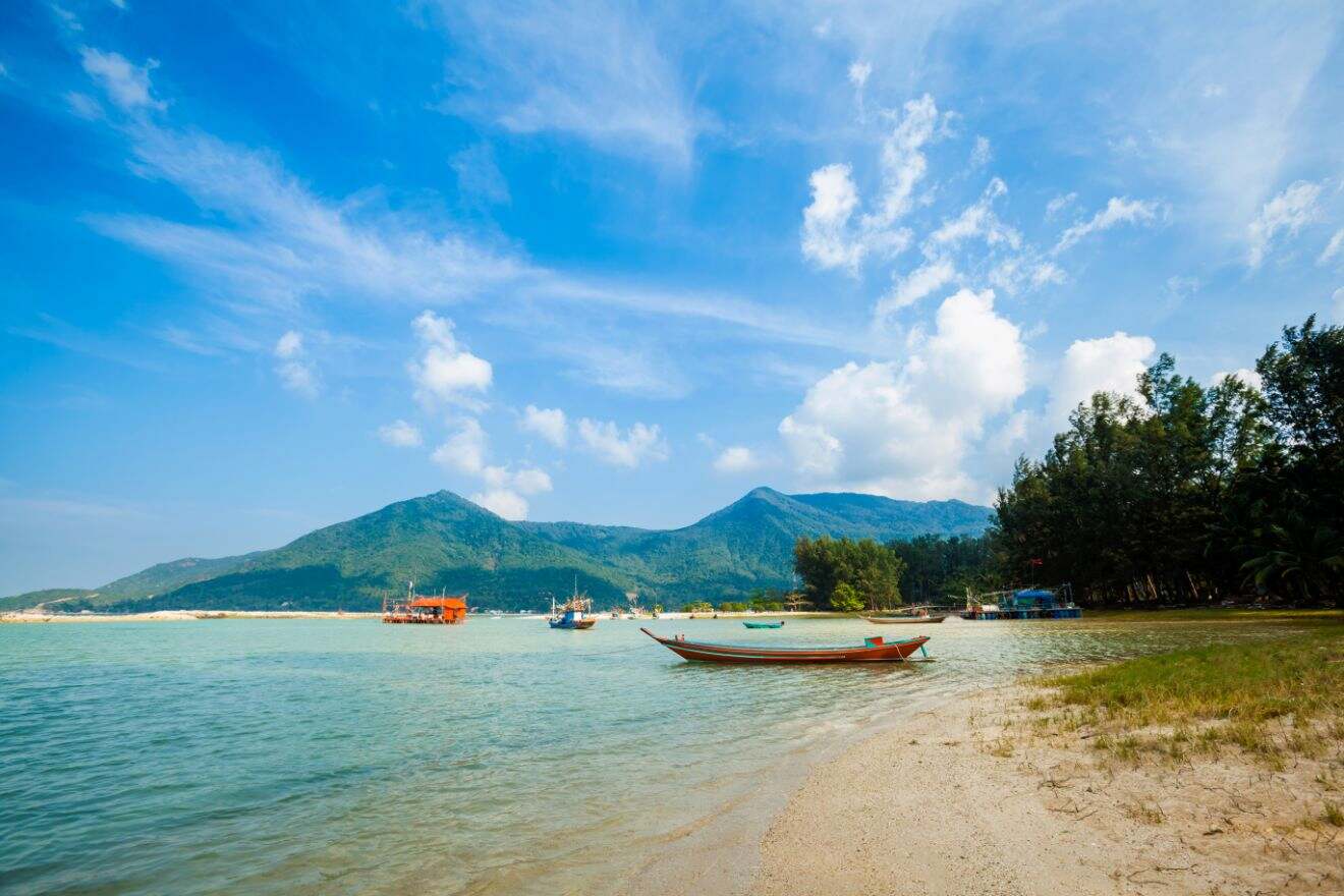 Scenic beachfront view in Chaloklum, Koh Phangan with a traditional wooden boat moored in shallow waters, with tropical greenery and mountains in the distance