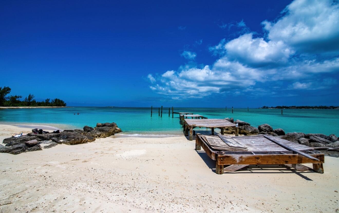 Serene beach scene in Nassau with a rustic wooden sunbed on pristine white sands, a small rock formation to the side, leading to a tranquil turquoise bay under a bright blue sky with scattered clouds.