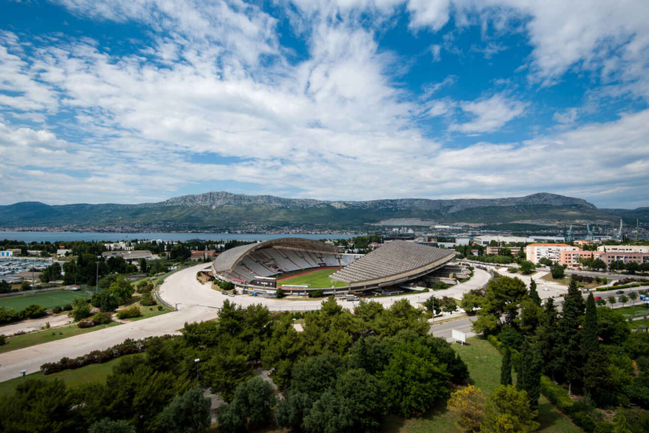 Wide-angle view of the Poljud Stadium in Split, Croatia, surrounded by lush greenery, with a backdrop of mountains and a clear blue sky with scattered clouds