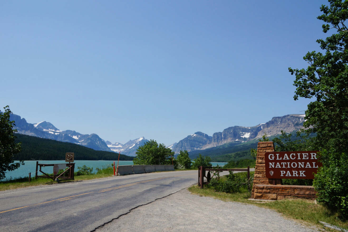 Entrance to Glacier National Park with a rustic wooden sign on a clear day, showcasing the park's towering mountain peaks and a turquoise lake