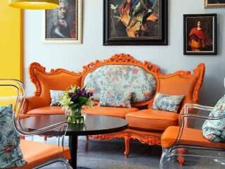 Vibrant living space featuring an ornate orange Victorian-style sofa set against a yellow wall adorned with eclectic paintings.