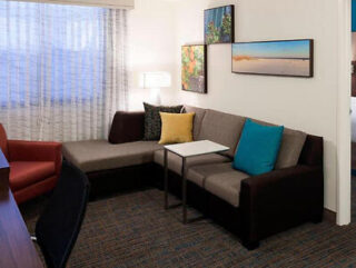 A homely living room corner in Residence Inn by Marriott featuring a sectional brown sofa with colorful cushions, a coffee table, and wall-mounted artwork