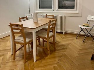 Simple dining space with a wooden table and chairs in a room with parquet flooring and a white radiator beneath a window