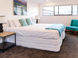 A neat, contemporary bedroom with a large king-size bed adorned with a teal throw, flanked by elegant nightstands and a comfortable turquoise chair