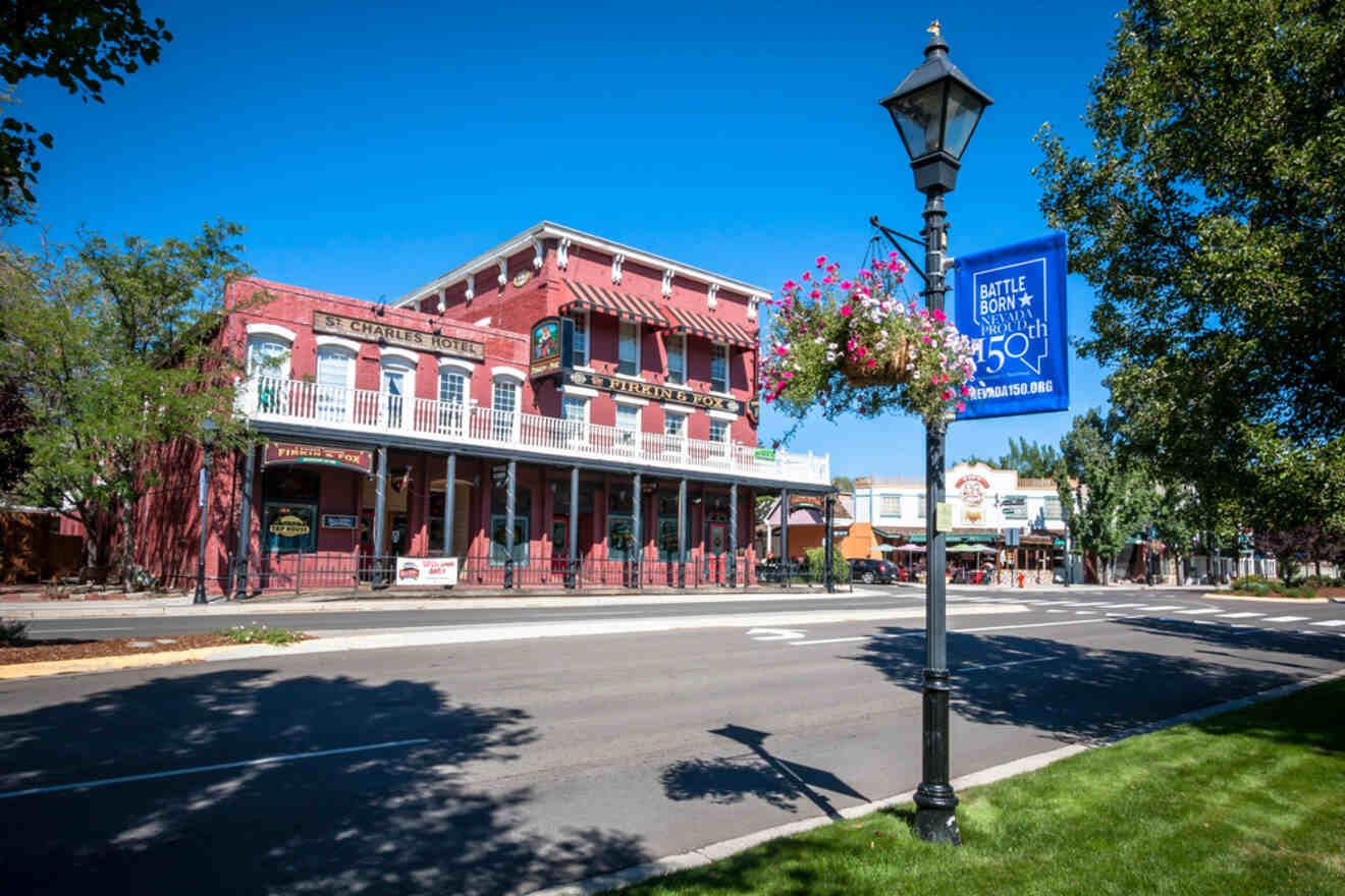 A historic red building with signage reading 'St. Charles Hotel' in Carson City, adorned with hanging flower baskets and a 'Battle Born Nevada 150' banner on a lamppost, under a bright blue sky on a sunny day.