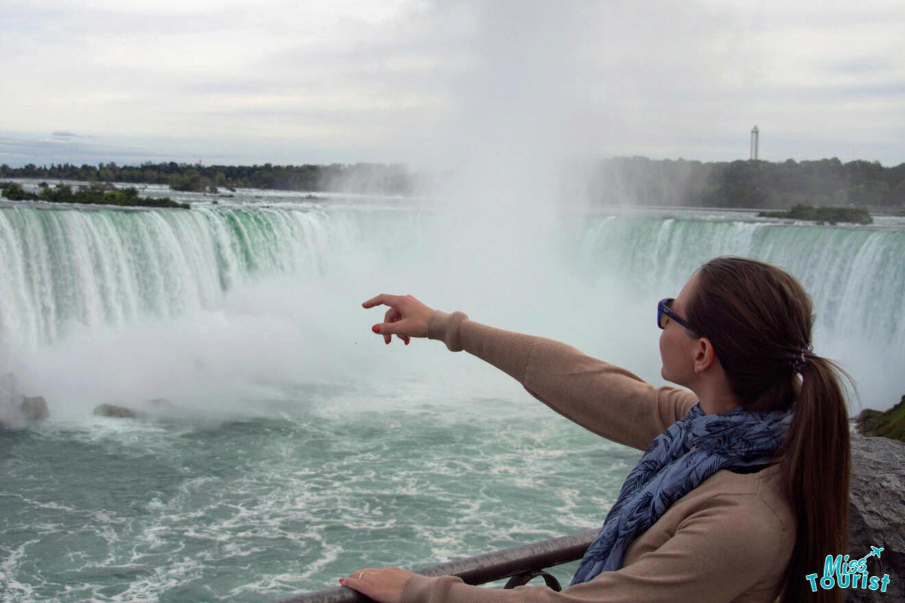 The writer of the post, Yulia, pointing at the mist of Niagara Falls, with the water's edge and lush greenery visible in the background