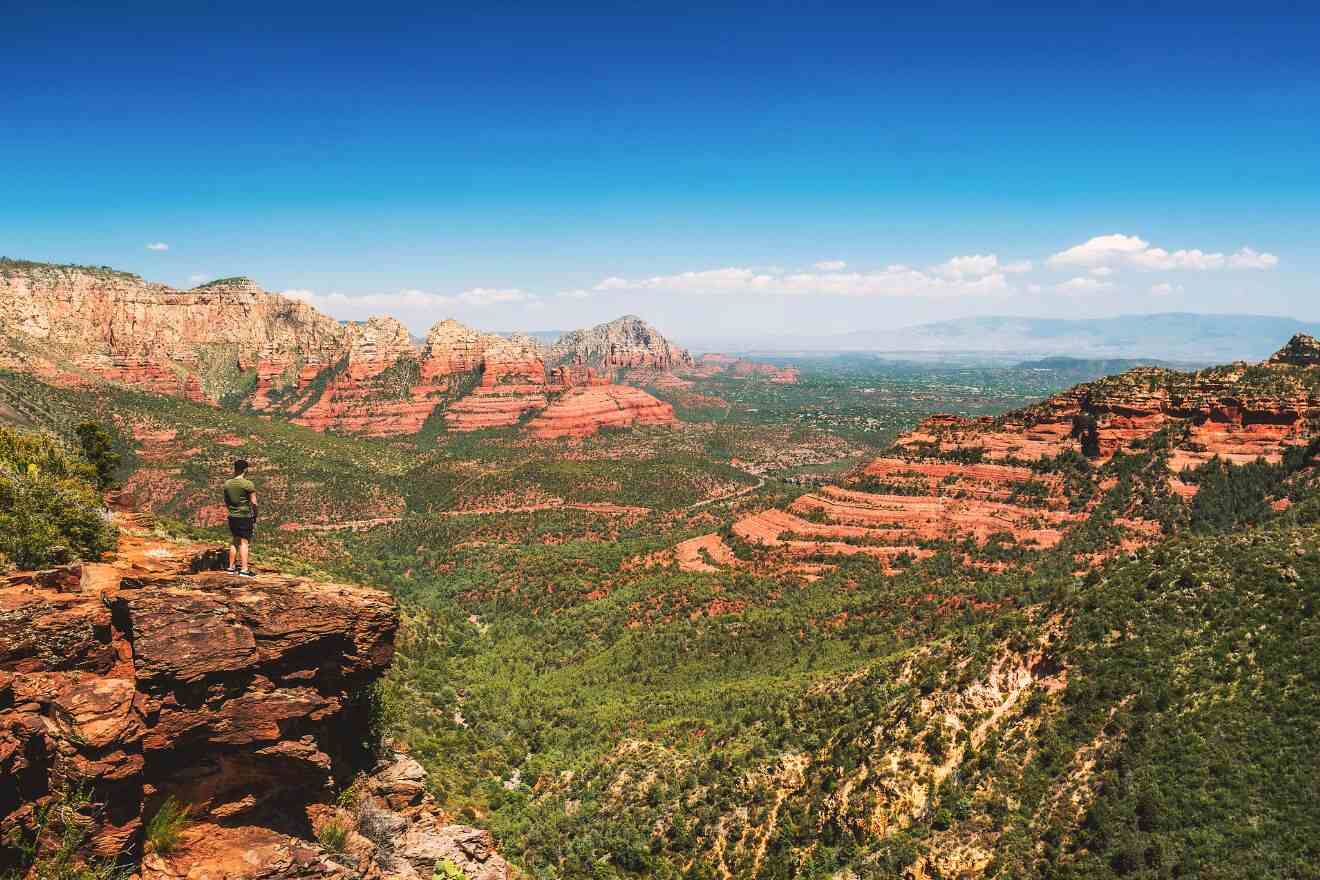 A hiker stands at the edge of a cliff overlooking the vast red rock canyons of Sedona under a clear blue sky